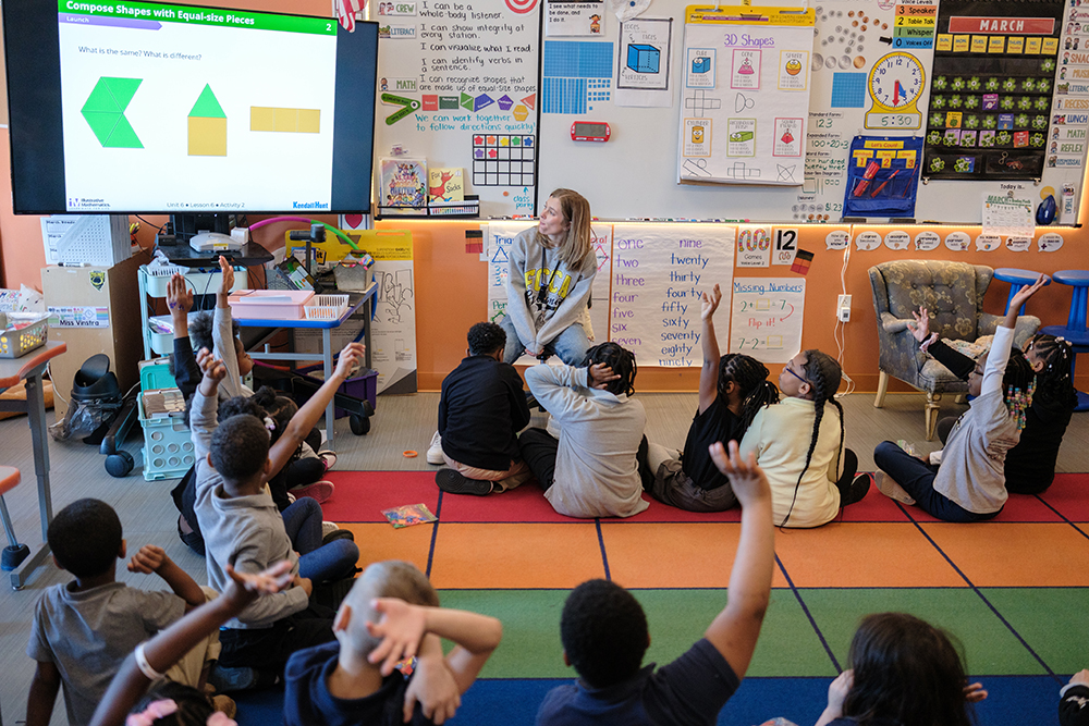 IMpact Research A classroom with student seated on the floor, all looking toward a tv screen with arms raised